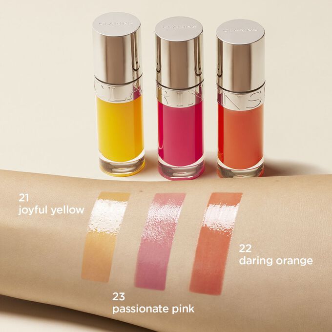 Présentation de la gamme d’huile lèvres Clarins dans 3 couleurs : jaune, rose et orange. Un échantillon de chacune a été apposé sur le bras d'une femme pour montrer le rendu du produit